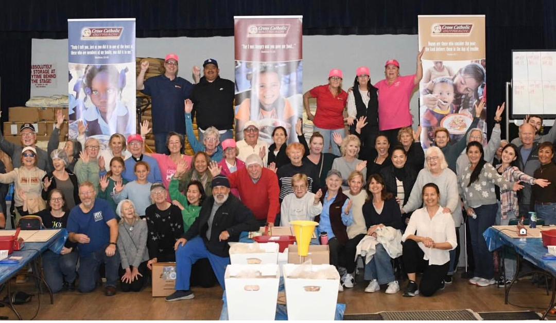 A large group of volunteers waving at the camera during a meal packing day, with charity banners behind them
